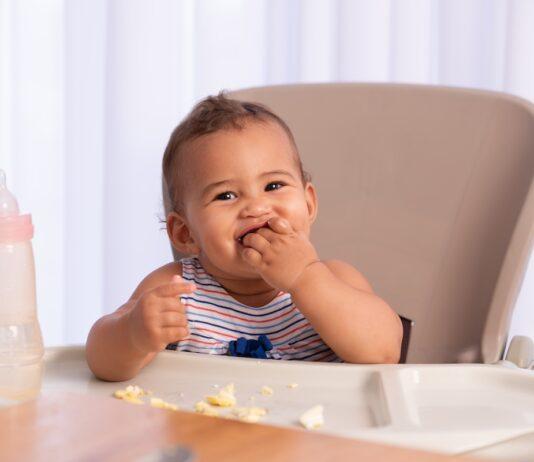 Cute baby child eating food, sitting on high chair.